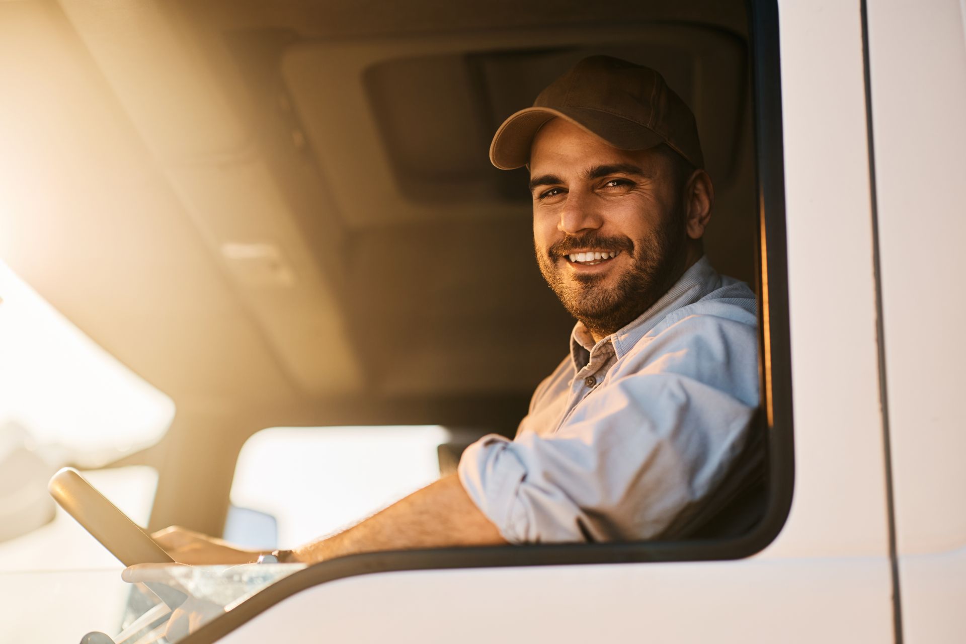 Smiling driver in a truck cab, holding steering wheel, wearing a cap. Sunlight shines in.