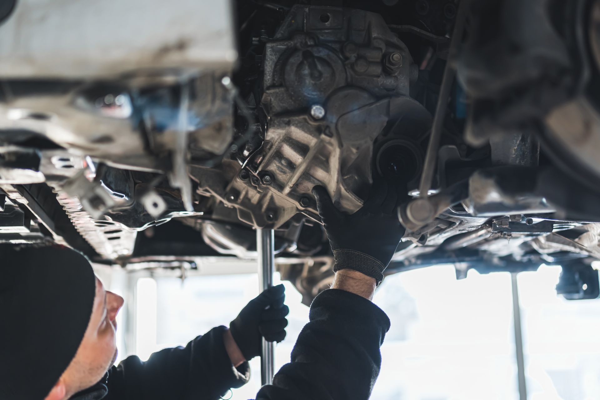 Mechanic working on a car from underneath, using a wrench. Black gloves and beanie visible.