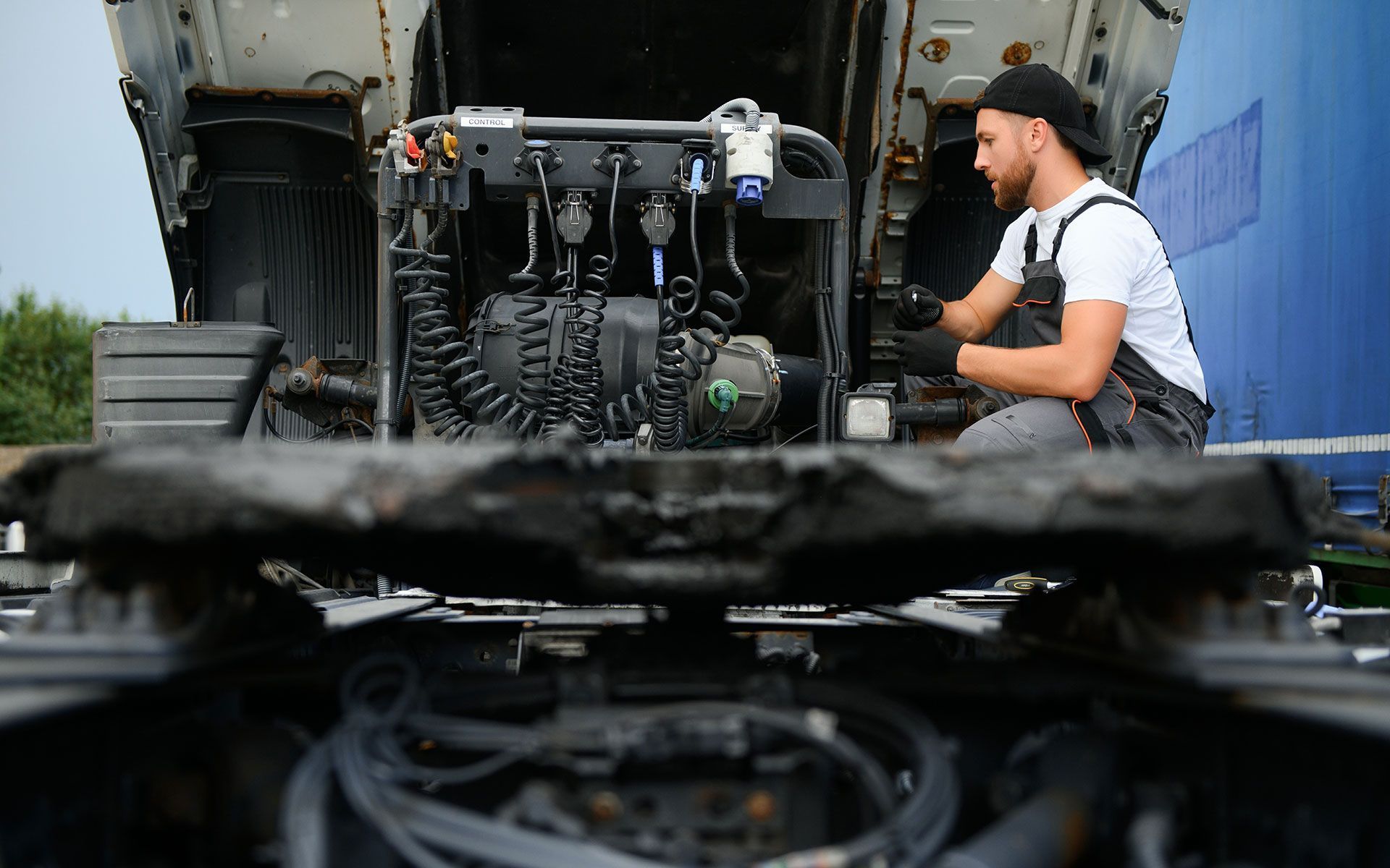 Mechanic works on a truck engine, wearing a cap and overalls.