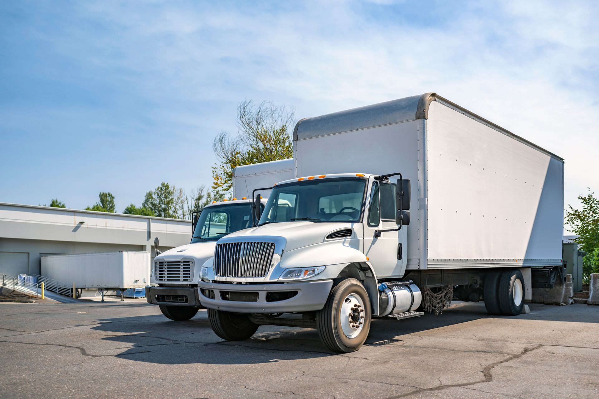 Two white box trucks parked outdoors on an asphalt lot under a blue sky.