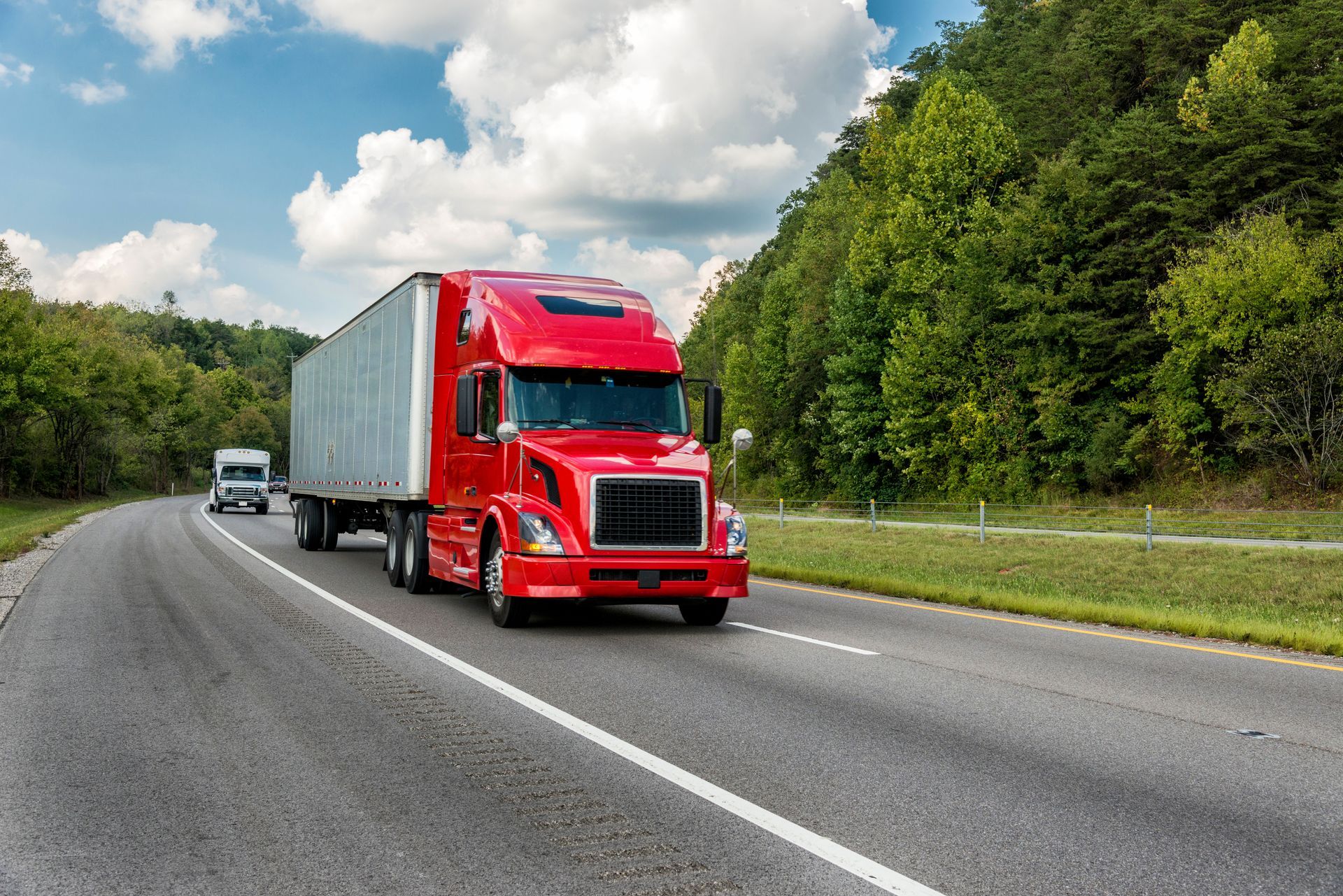 Red semi-truck on highway, green trees in background, sunny day.