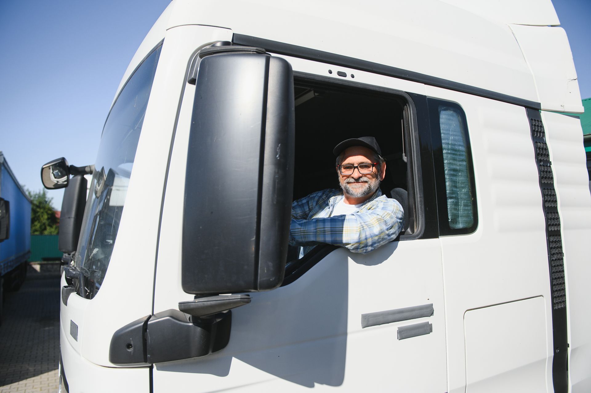 Truck driver in a white cab, smiling and looking out window, arms crossed.