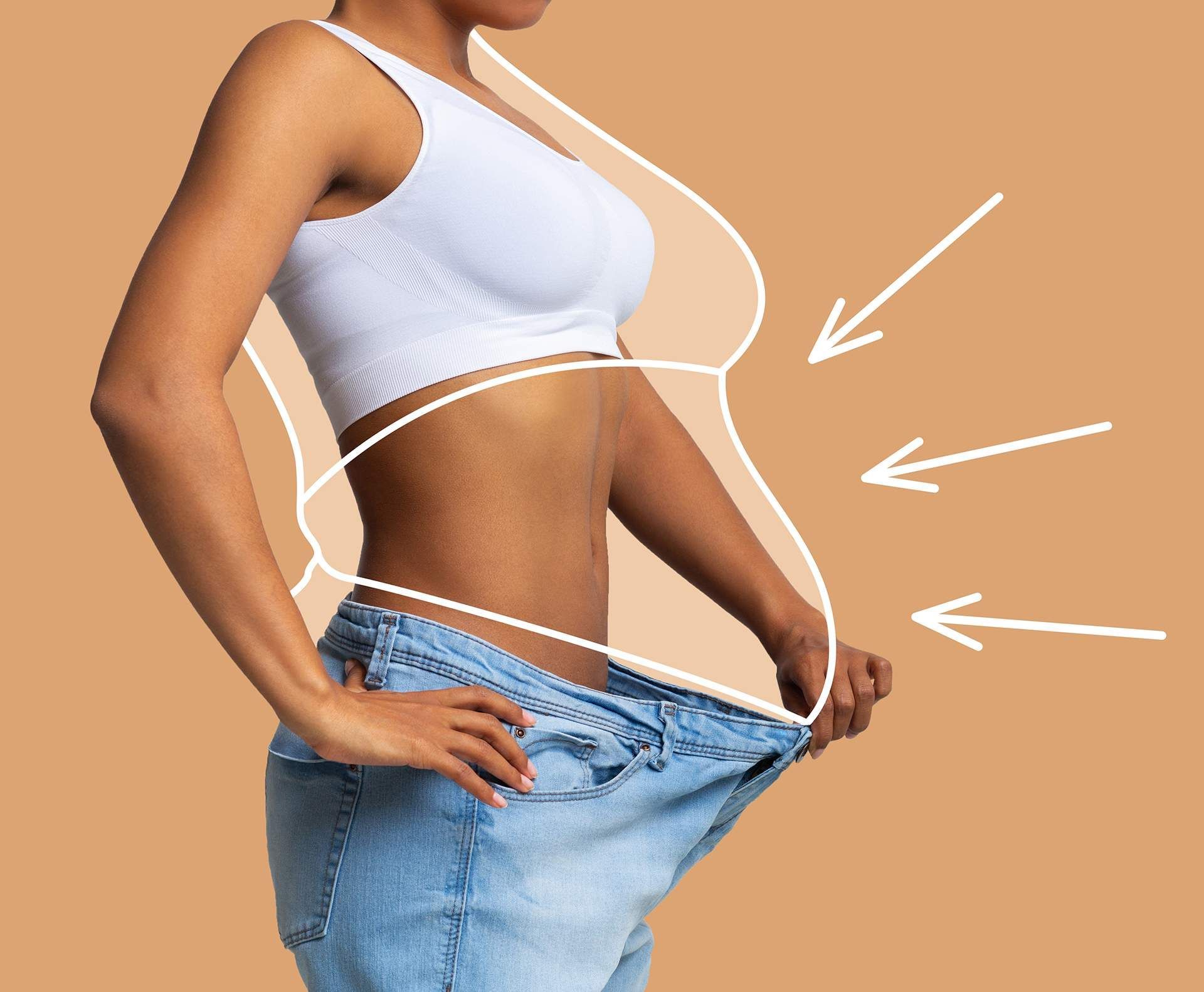 Woman in oversized jeans showing weight loss, wearing a white top, tan background.