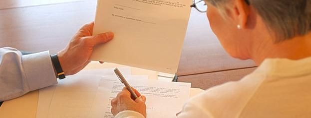 Dos personas revisando documentos; una persona sosteniendo un papel, otra escribiendo con un bolígrafo.