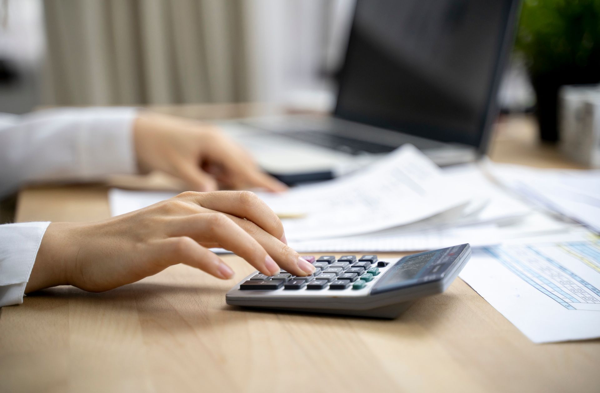 Hands using a calculator on a desk with documents, a laptop, and a plant, suggesting financial work.