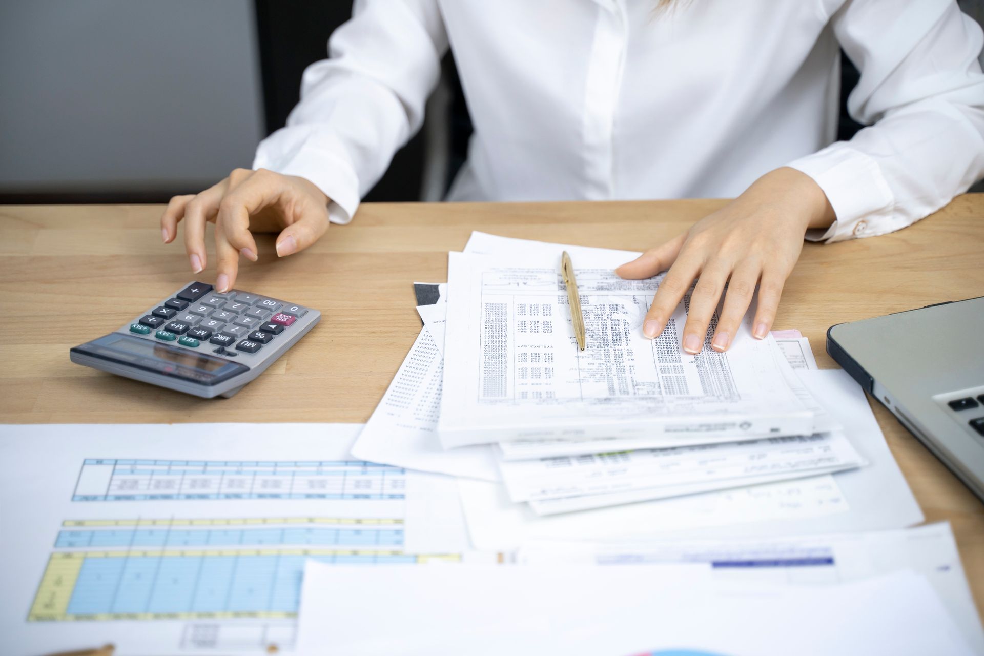 A person at a wooden desk calculates with a calculator, reviewing documents and a laptop.