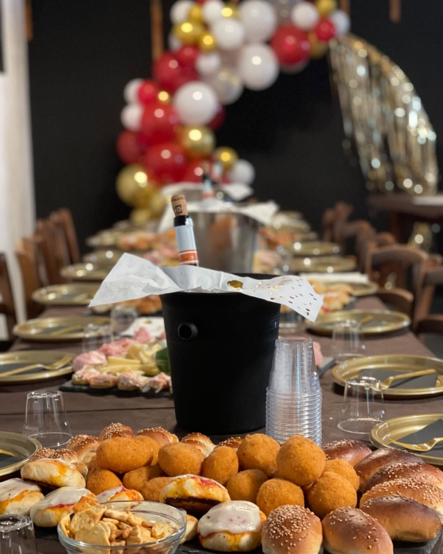 Tavolo da festa apparecchiato con cibo, bevande, piatti e arco di palloncini. Secchiello nero per bottiglie.