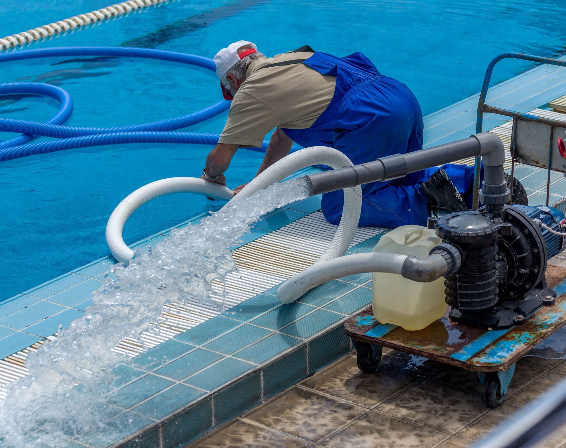 Man in blue overalls uses a pump to drain a swimming pool, water flowing onto tiled deck.