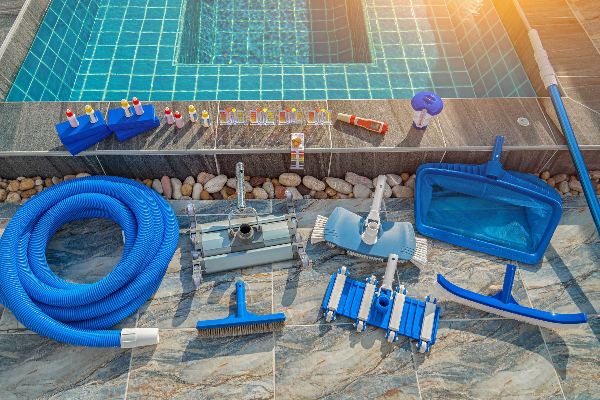 Pool cleaning tools arranged on a tiled surface beside a swimming pool; includes hose, brushes, nets, and chemicals.