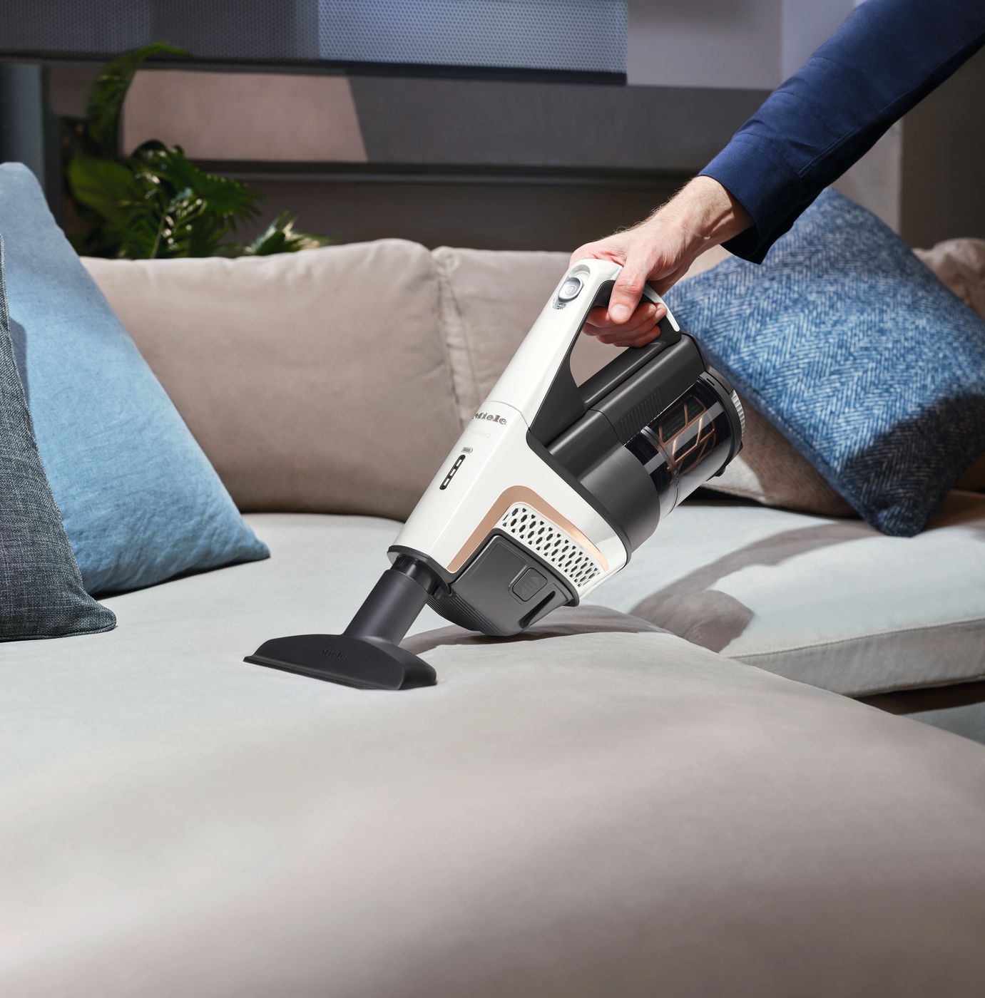 A person vacuuming a light-colored sofa with a white and black handheld vacuum.