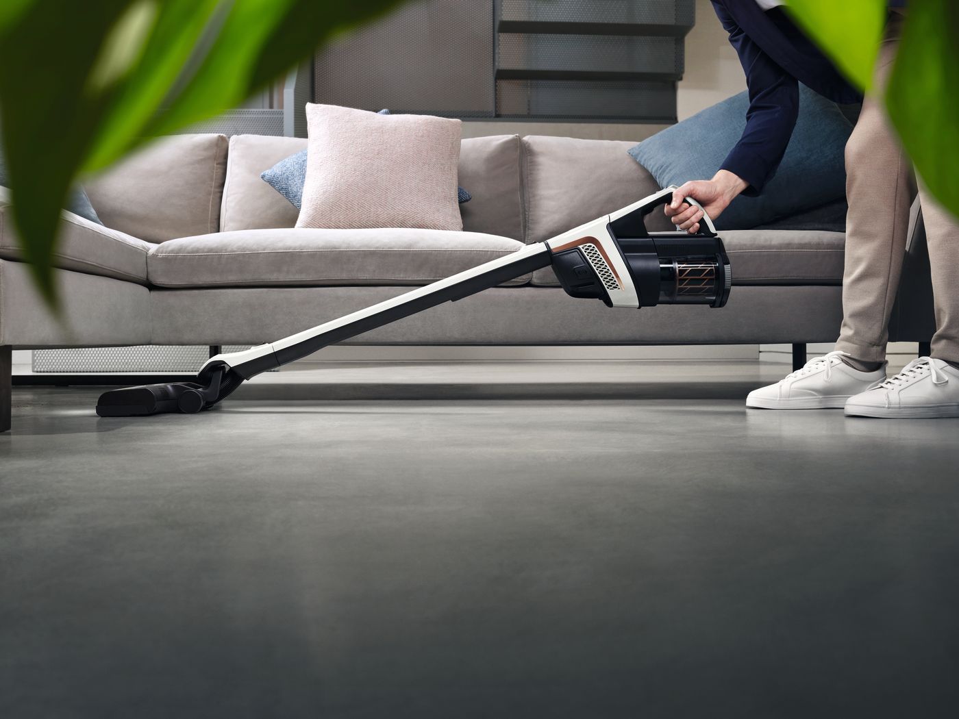 Person vacuuming a grey floor with a cordless vacuum cleaner near a couch.