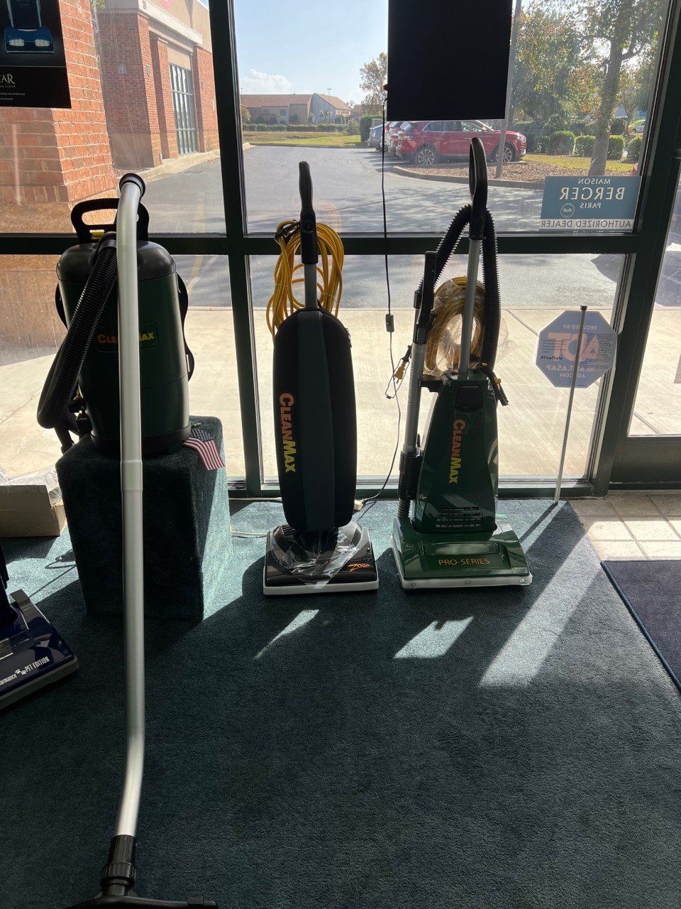 Three vacuum cleaners in a storefront window. Green and black machines on carpet, sunlight, and a view of the outside.