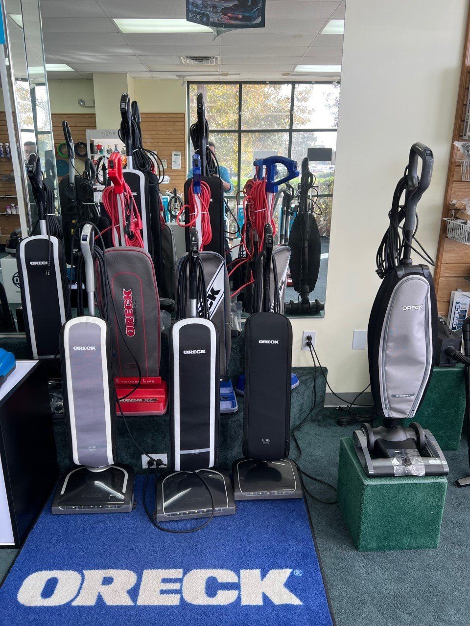 Vacuum cleaners in a store, many uprights. Oreck logo rug.