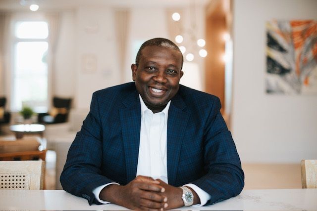 A man in a suit is sitting at a table with his hands folded and smiling.
