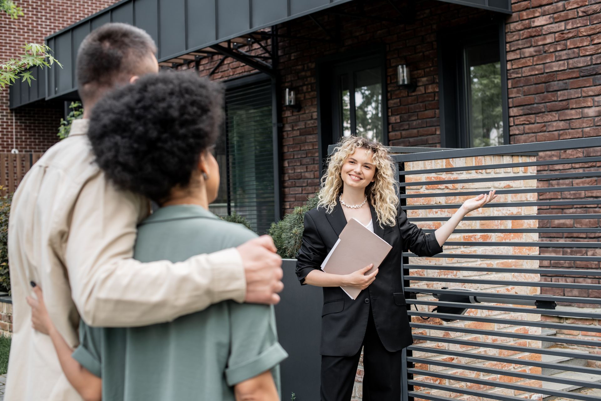 A woman is standing in front of a brick house talking to a couple.