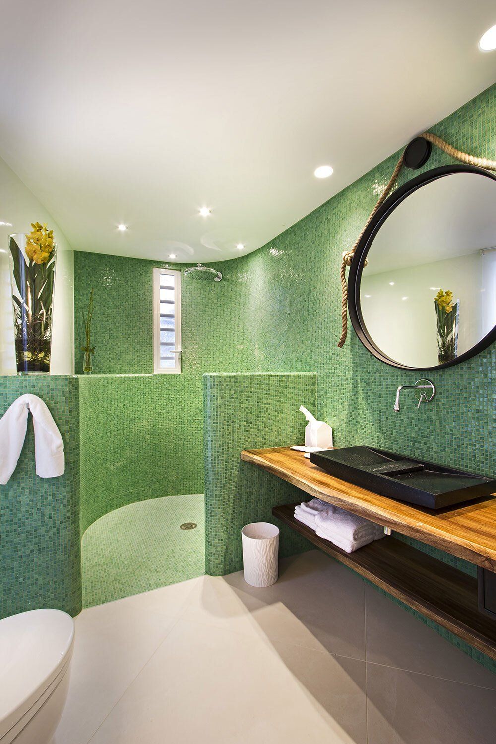 A bathroom with green tiles , a toilet , sink and mirror.