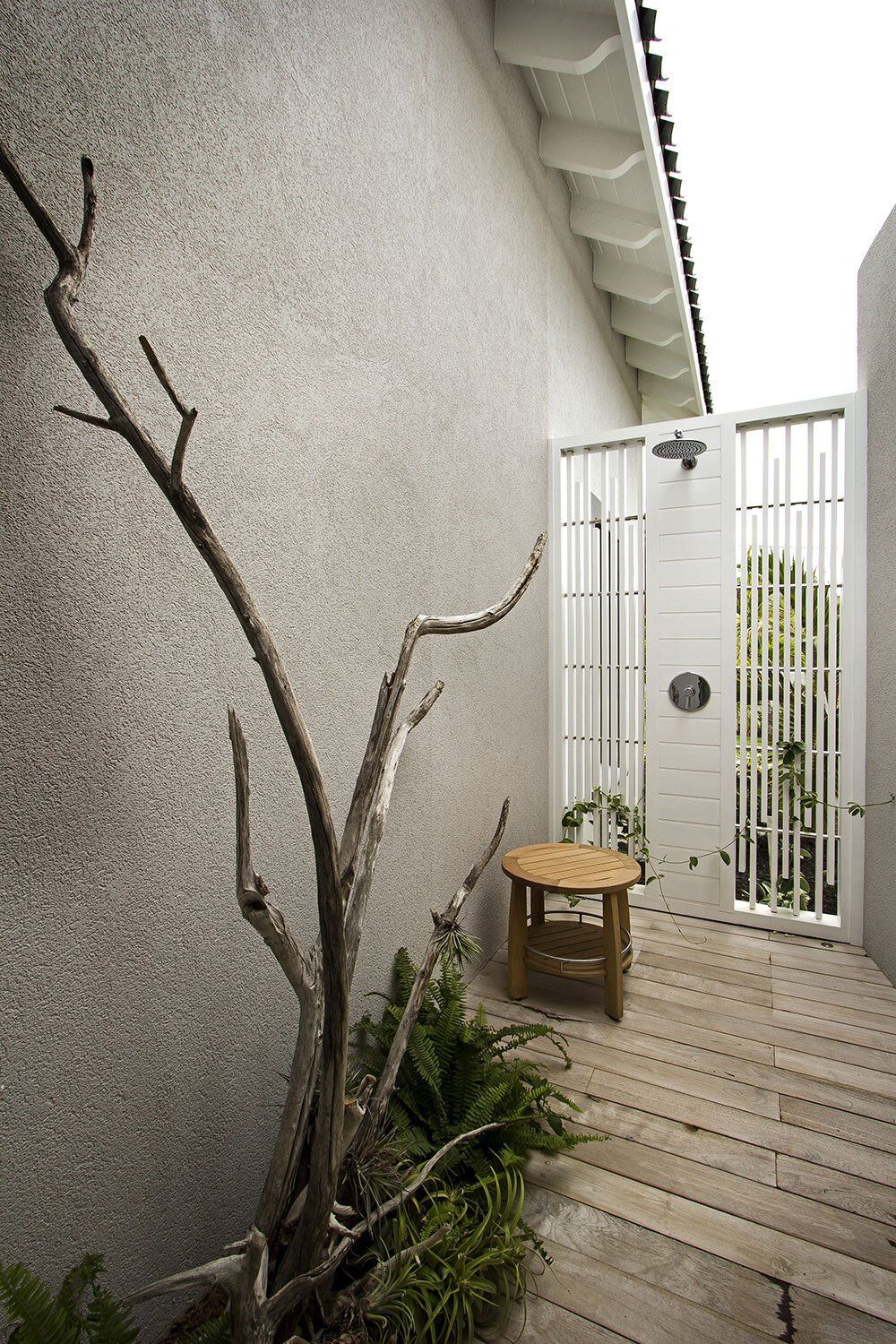 A wooden stool is sitting on a wooden deck next to a white wall.