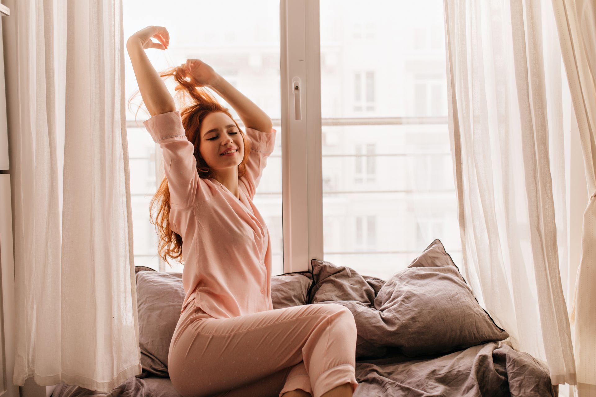 A woman is sitting on a bed stretching her arms in front of a window.