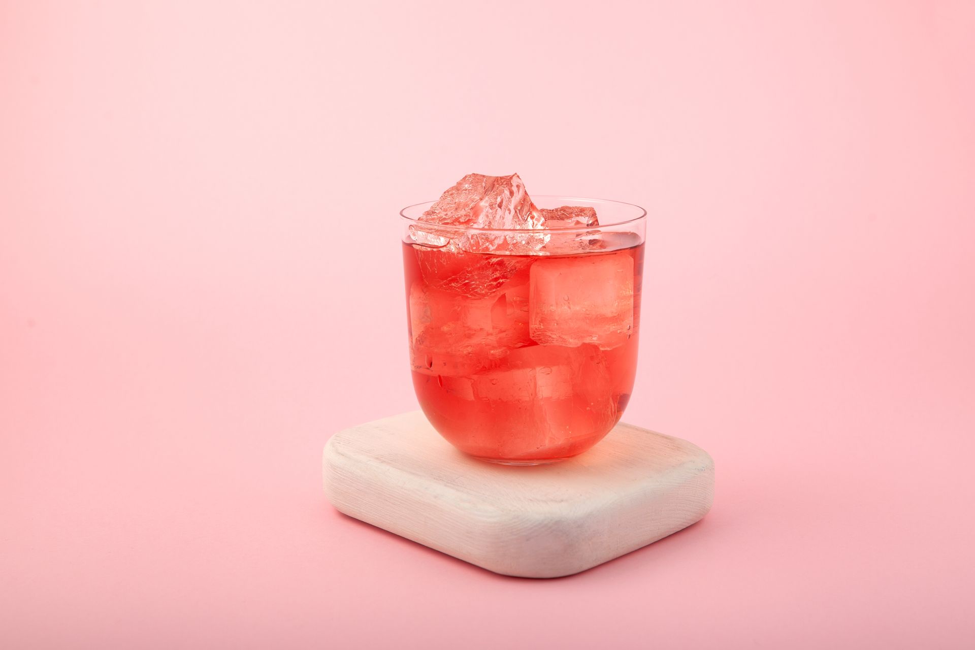 A glass of red liquid with ice cubes on a pink background.