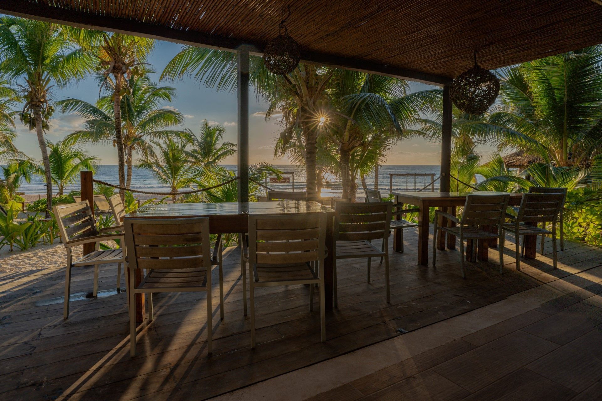 Wooden tables and chairs on a beachfront patio under a thatched roof, with palm trees and ocean in the background.
