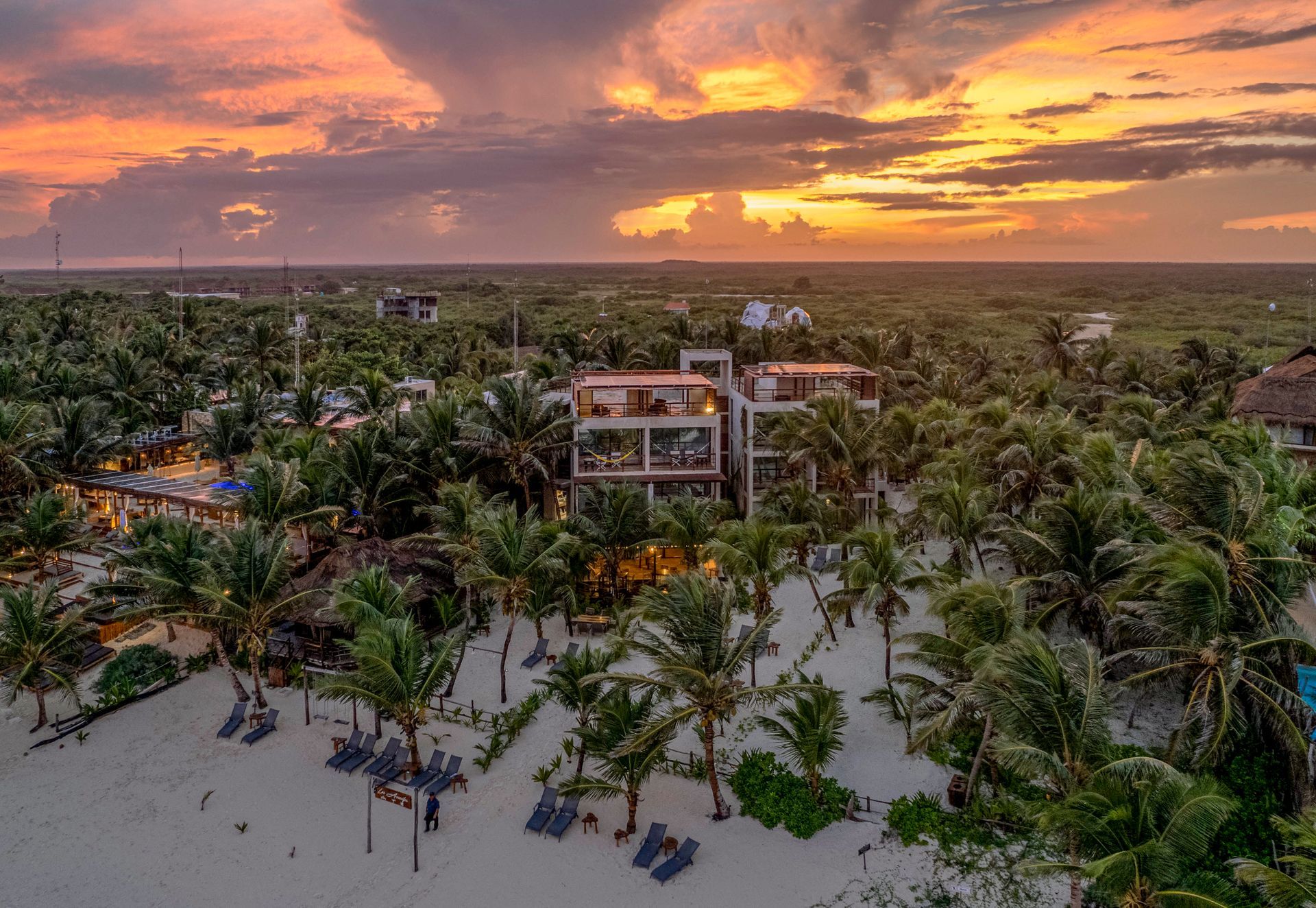 Aerial view of beach with palm trees and a modern building, sunset sky.