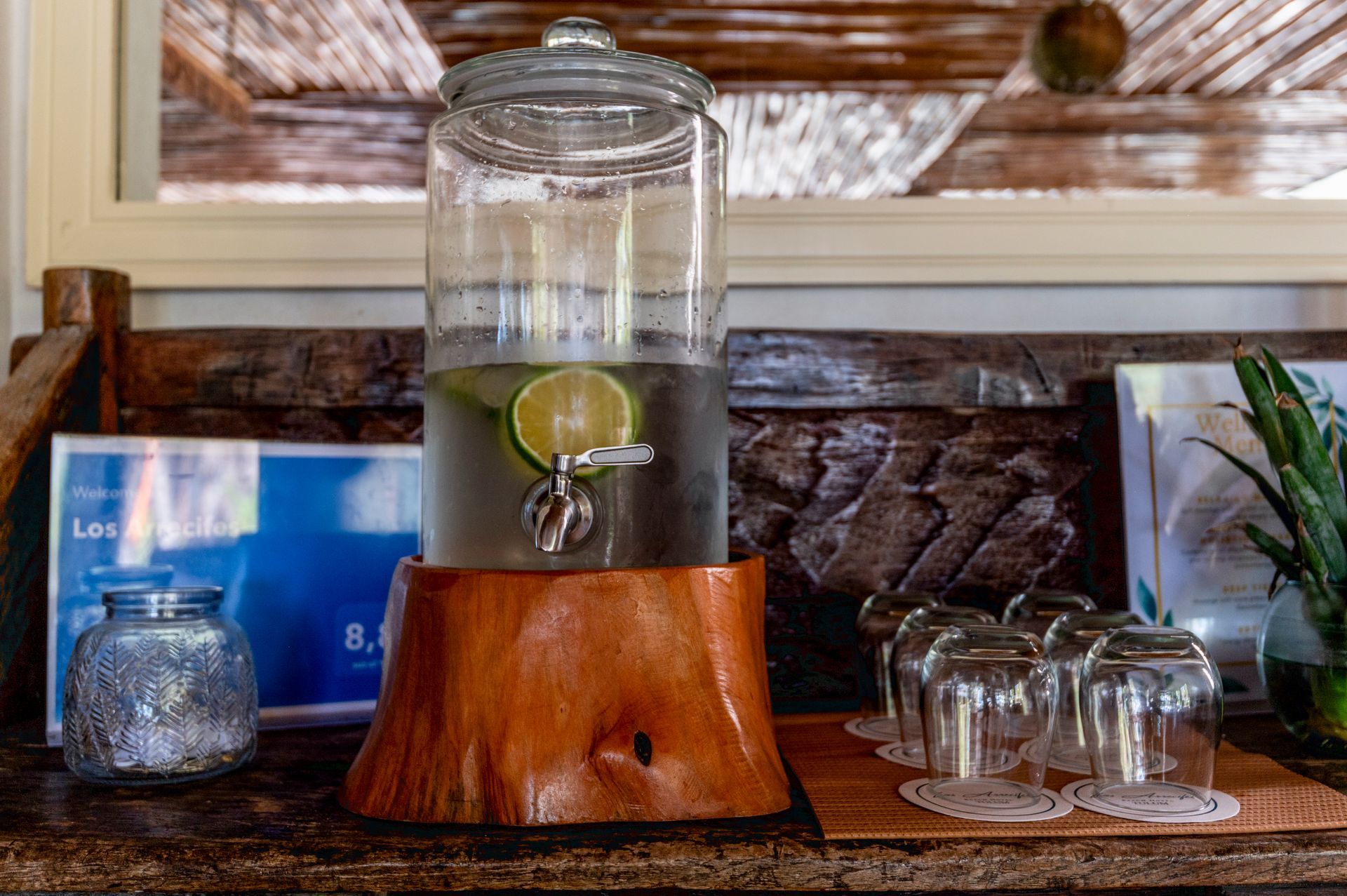 Water dispenser with lime slices, wooden base, glasses, and a jar on a wooden counter.
