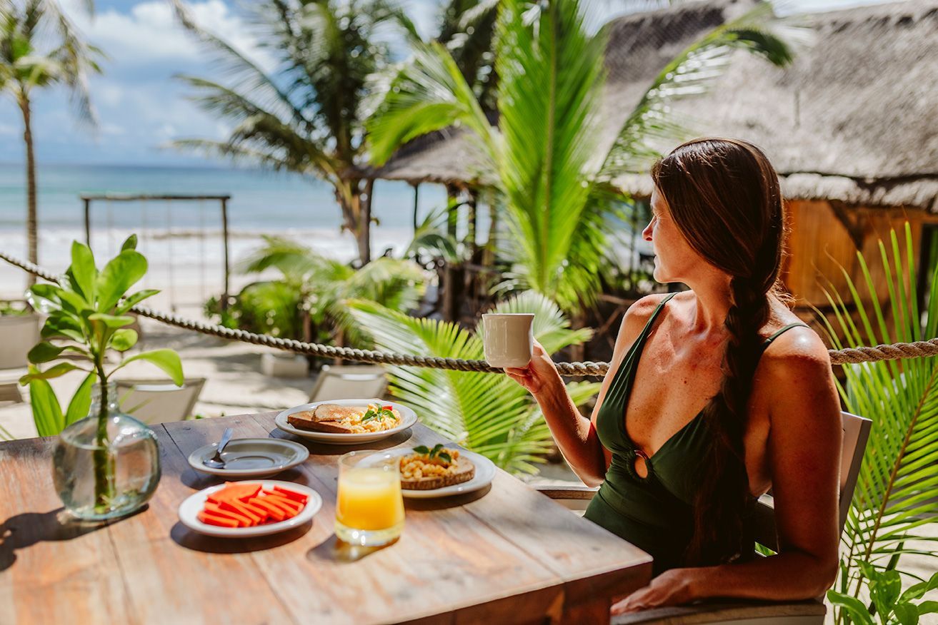 Woman in green swimsuit at beachside table, looking at the ocean, with breakfast and a coffee.