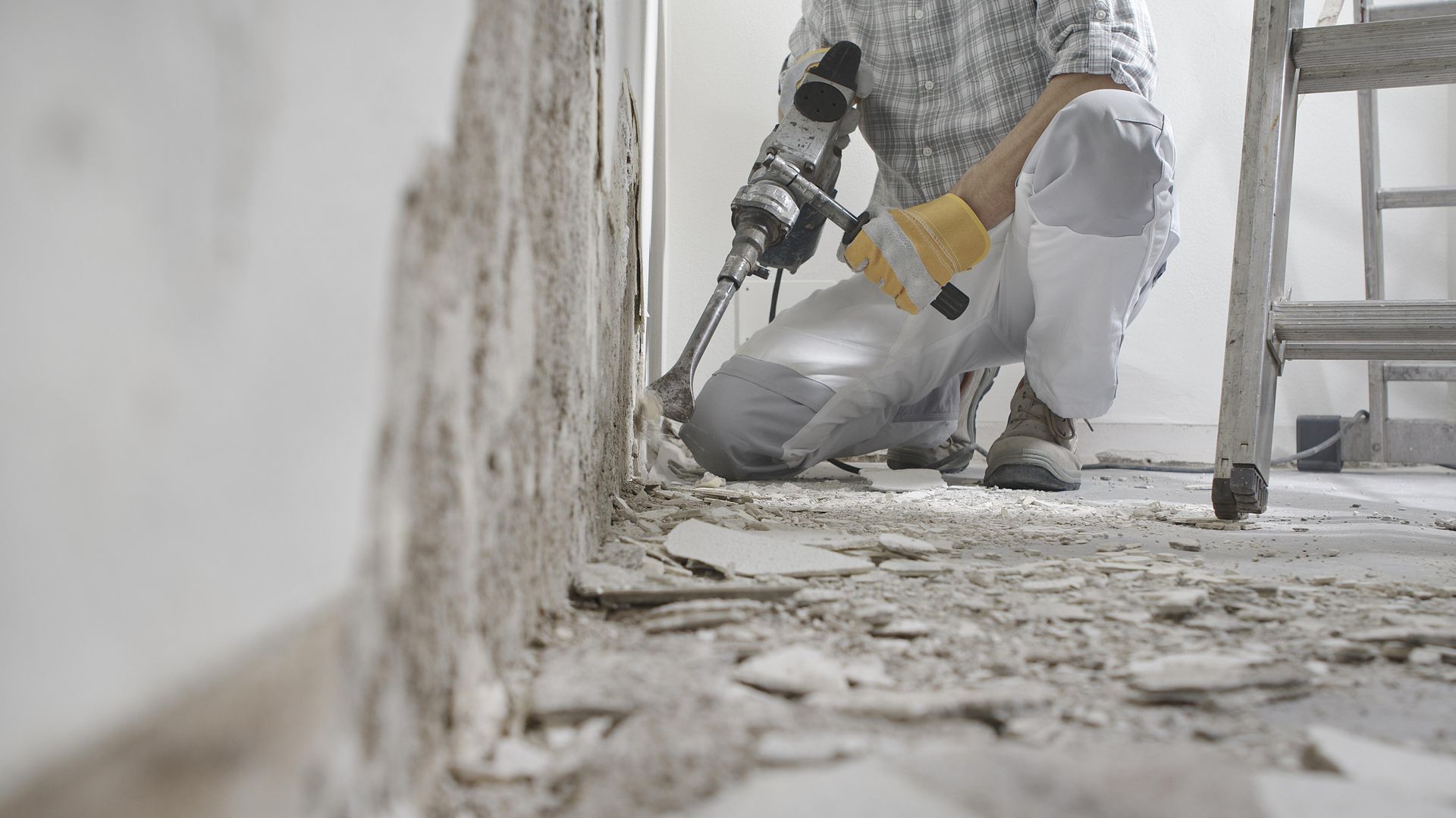 Person kneeling, using a demolition hammer to remove plaster from a wall in a room.