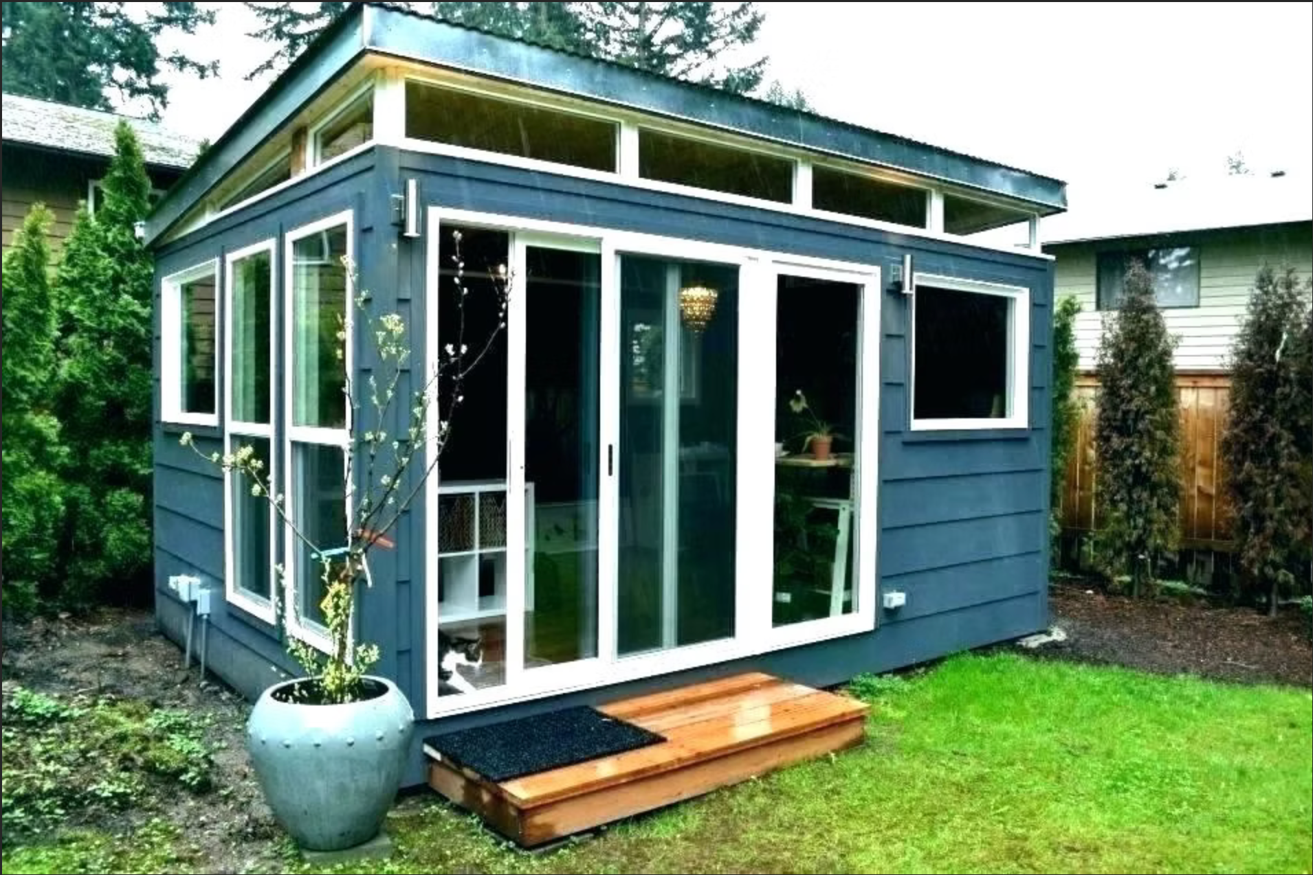 Small blue backyard shed with glass doors and windows, wooden steps, and a grassy yard.