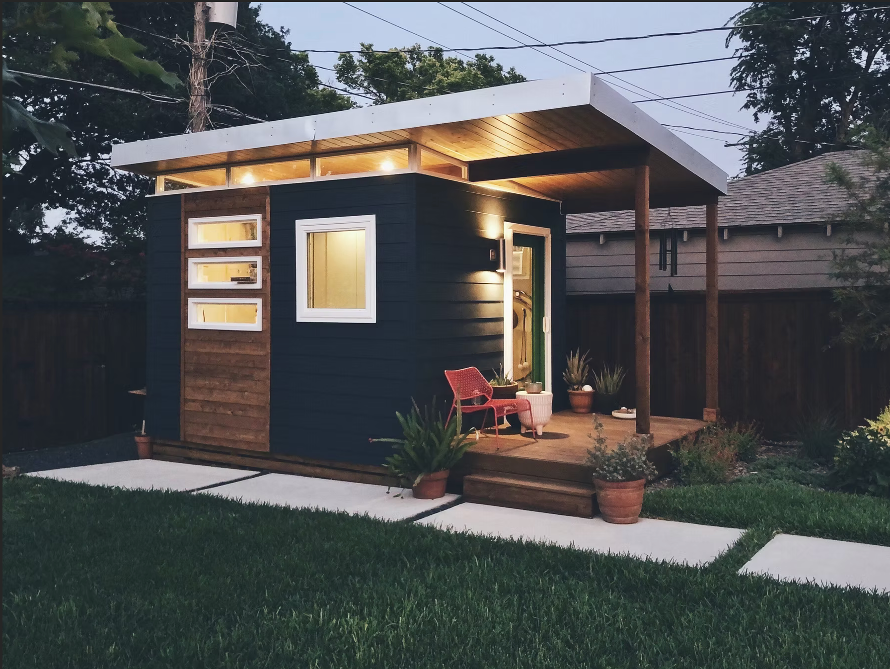 Small dark blue modern backyard shed with a wooden deck and walkway.