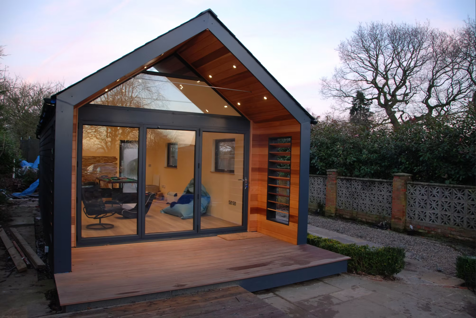 Modern backyard cabin with glass front and wooden deck, set against a background of trees and gravel.