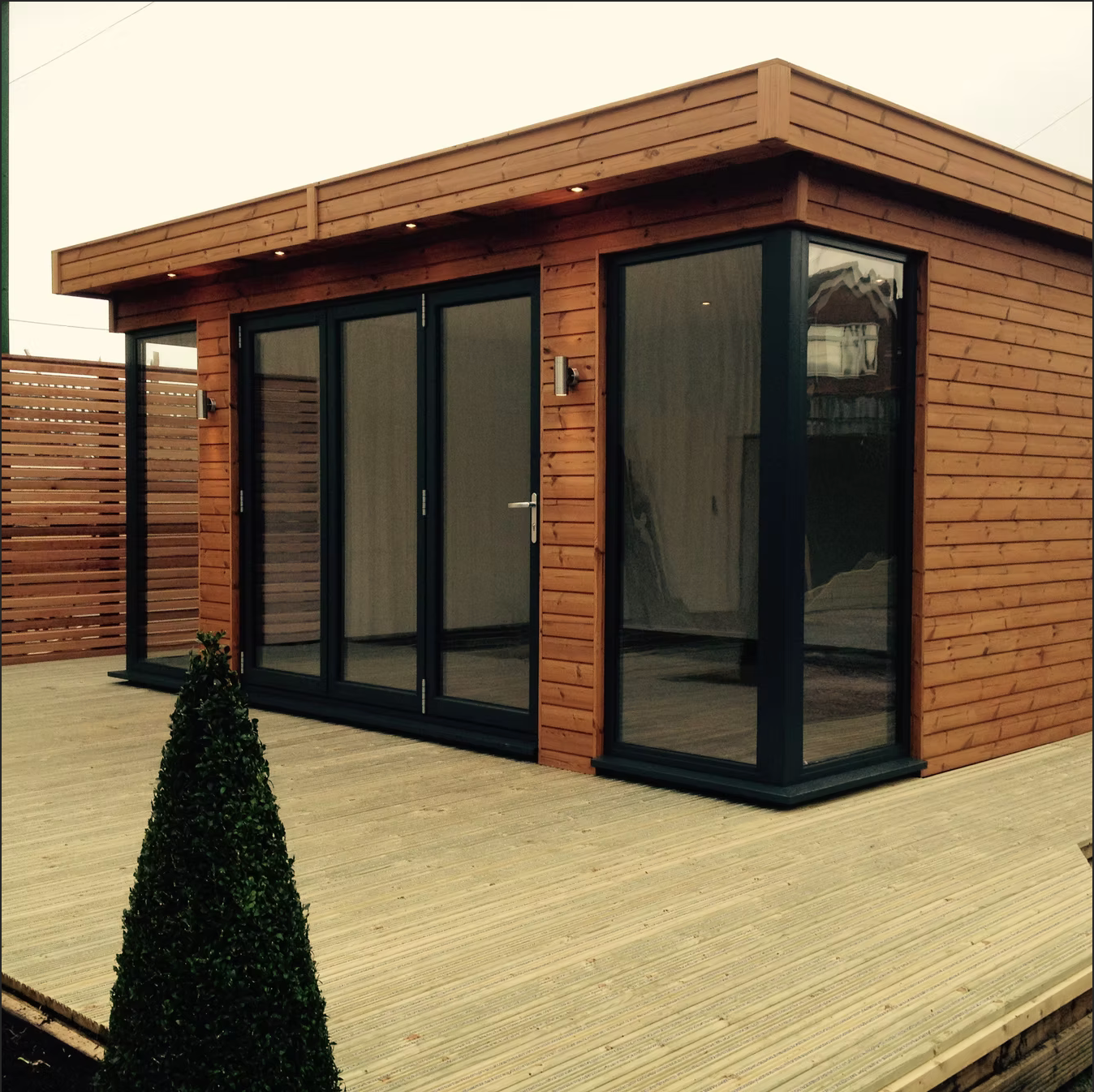 Wooden garden office with large glass windows and doors on a deck, brown siding.
