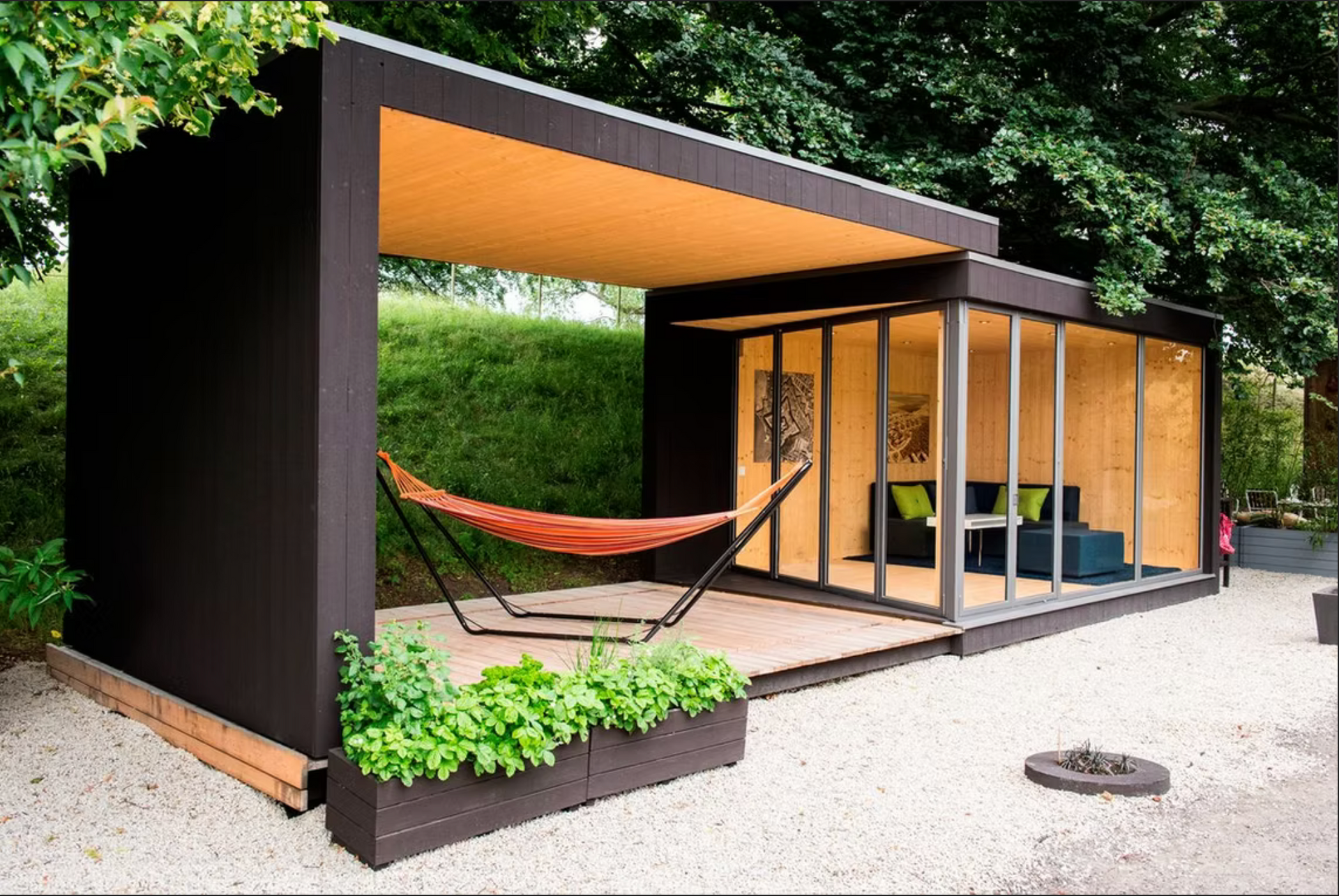Modern outdoor structure with a hammock, seating area, and sliding glass doors. Brown exterior, gravel ground, and greenery.