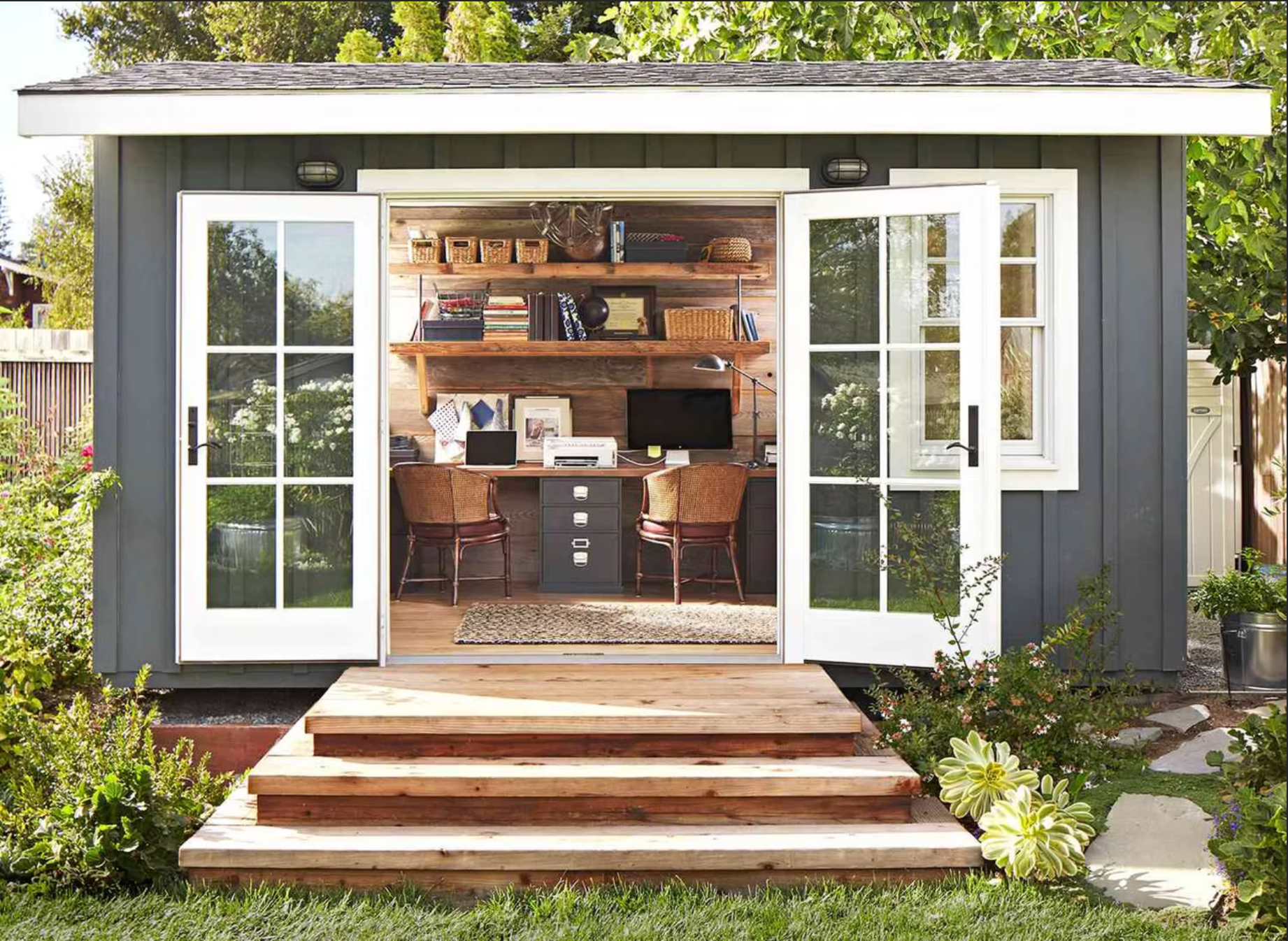 Gray shed with open glass doors, wooden steps, and an interior workspace with shelves and a desk.