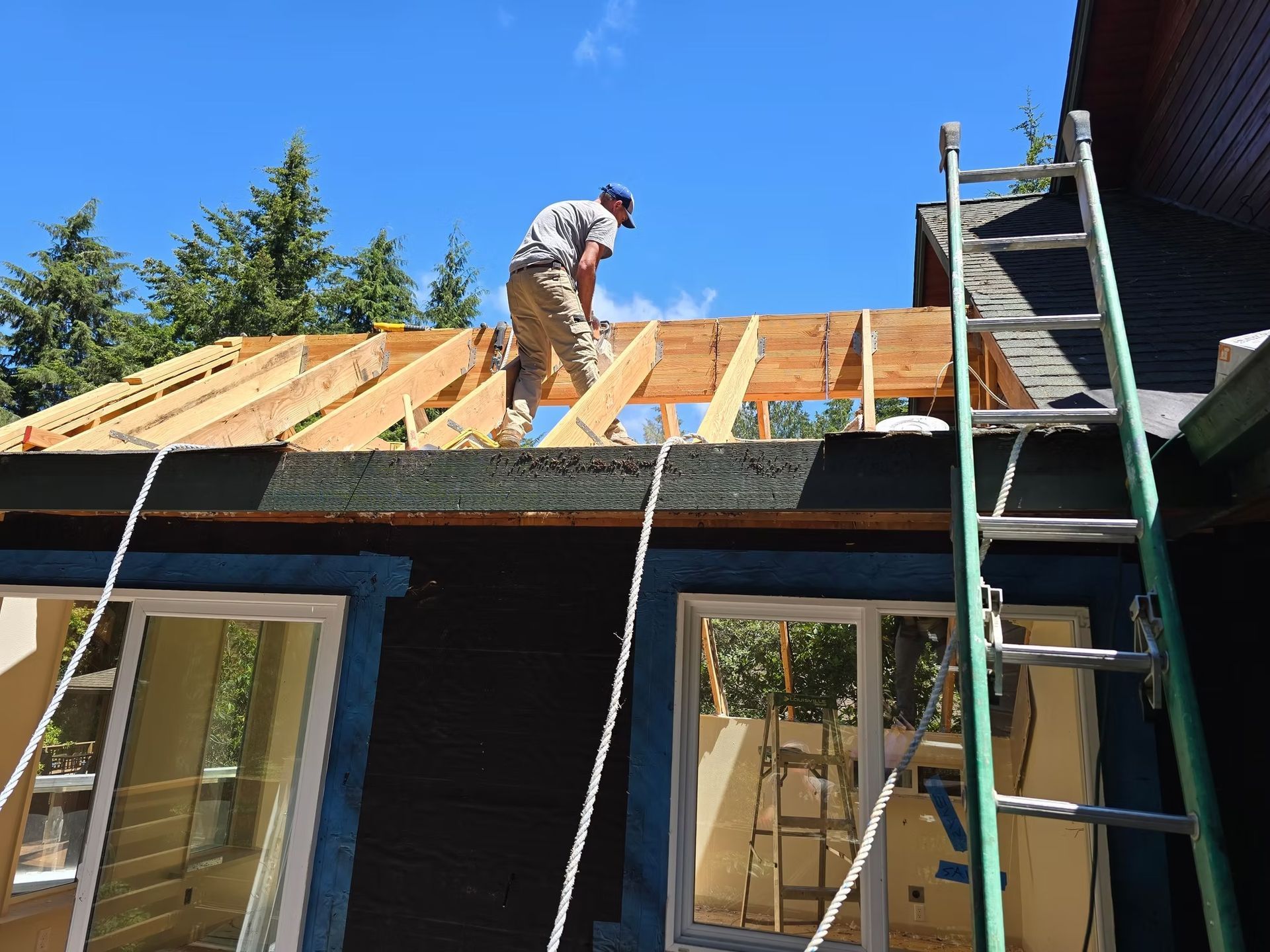 Man on rooftop building frame, blue house with windows, ladder, sunny day.