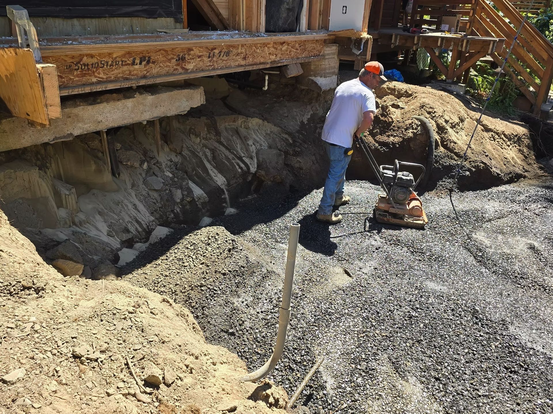 Man compacting gravel under a building with a compactor. Exterior construction site with wooden structure.