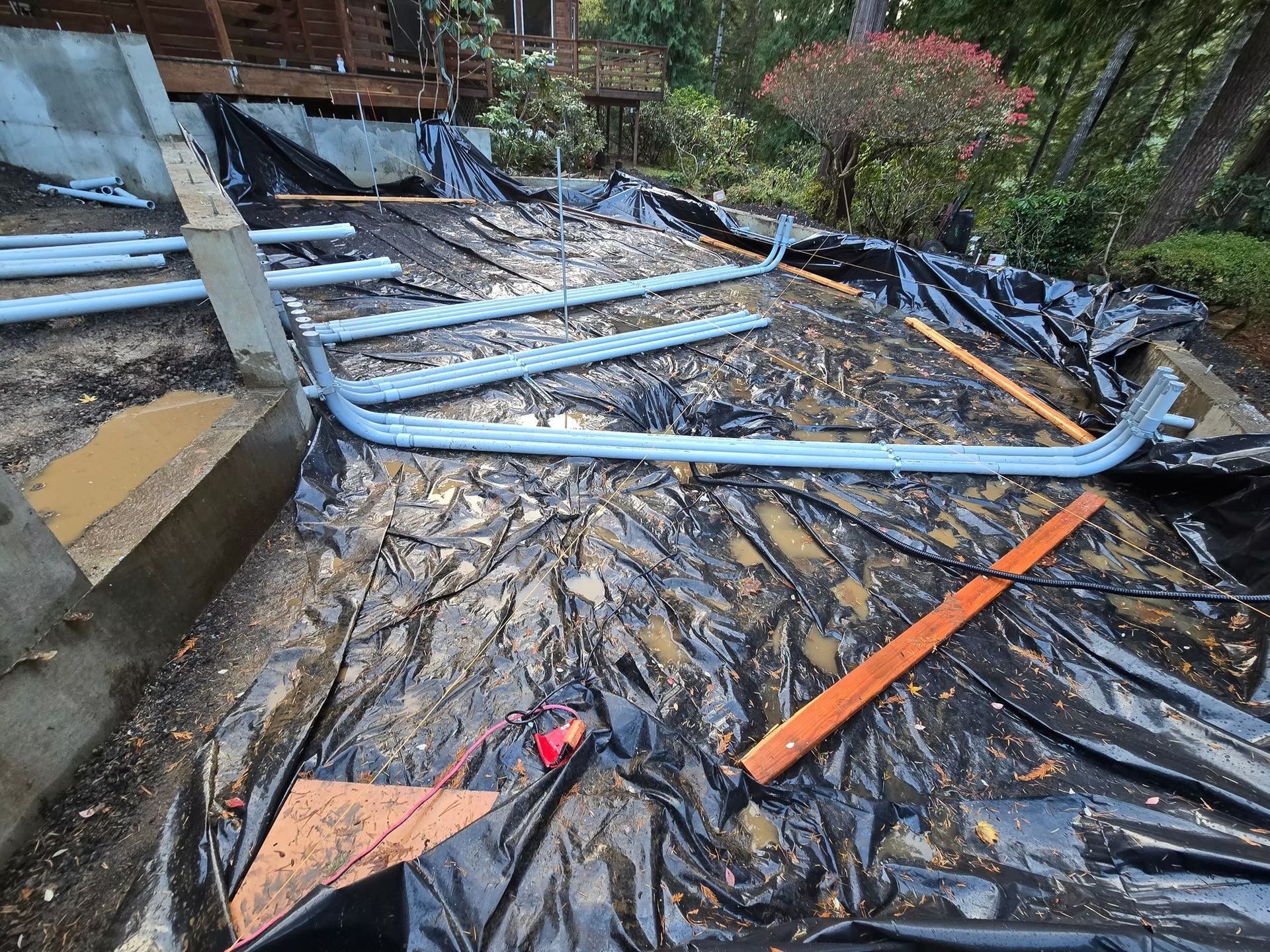Construction site with black tarp, gray pipes, and wooden beams on a sloped outdoor area.