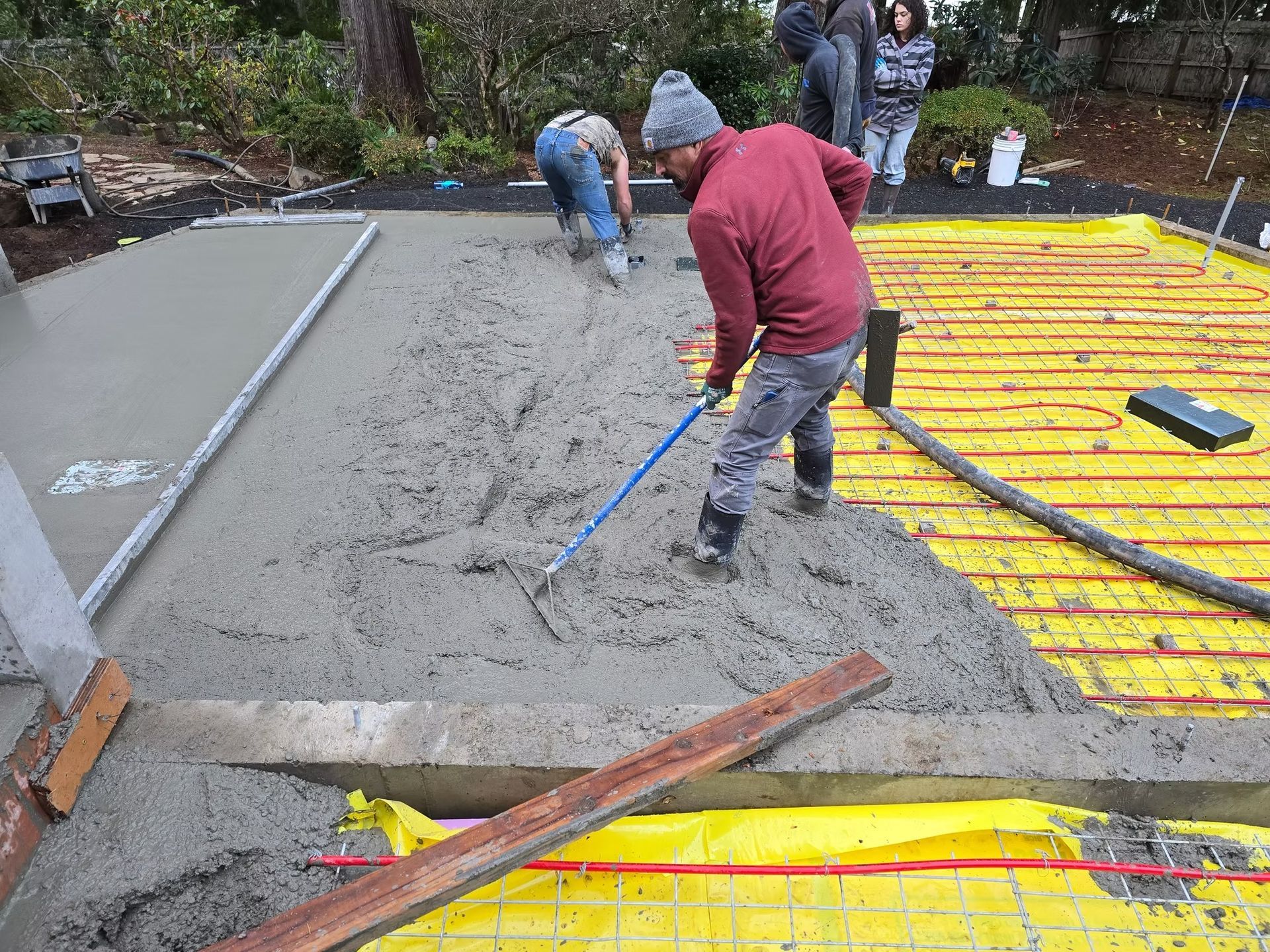 Workers pouring and smoothing wet concrete over yellow tubing on a concrete slab in an outdoor setting.