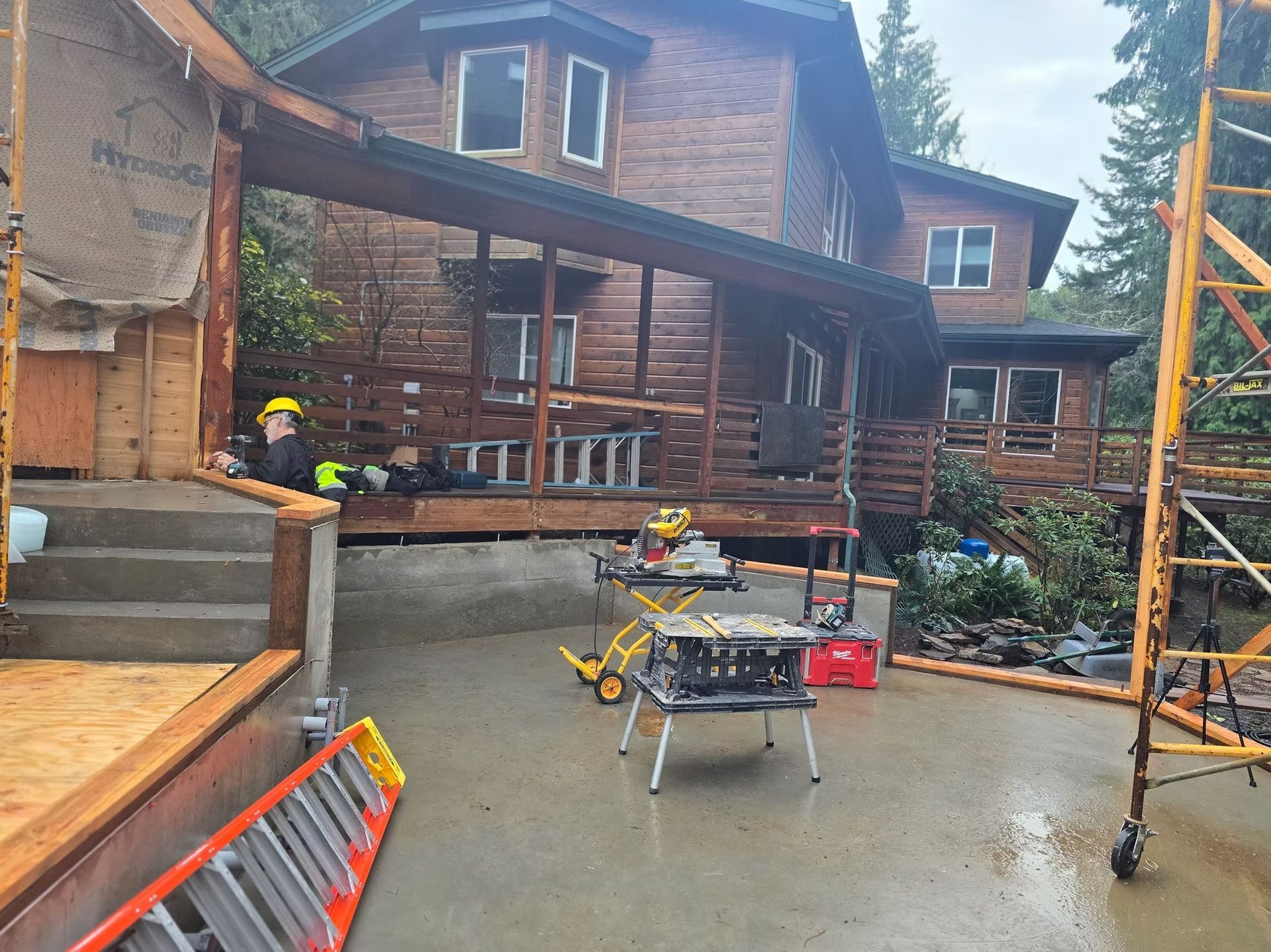 Construction site: House with wooden siding, porch, and a person in a hard hat; tools and scaffolding present.