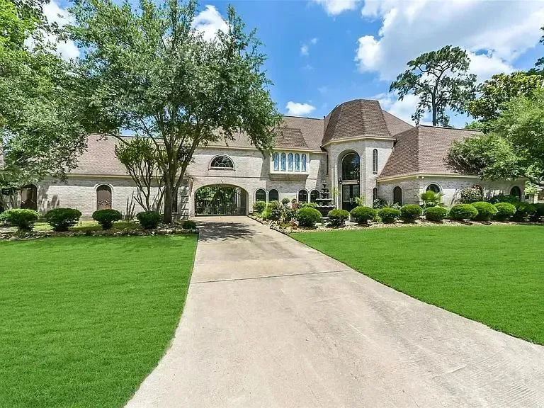 A two-story brick mansion with a large front lawn, an arched driveway, and a shingled roof under a blue, cloudy sky.