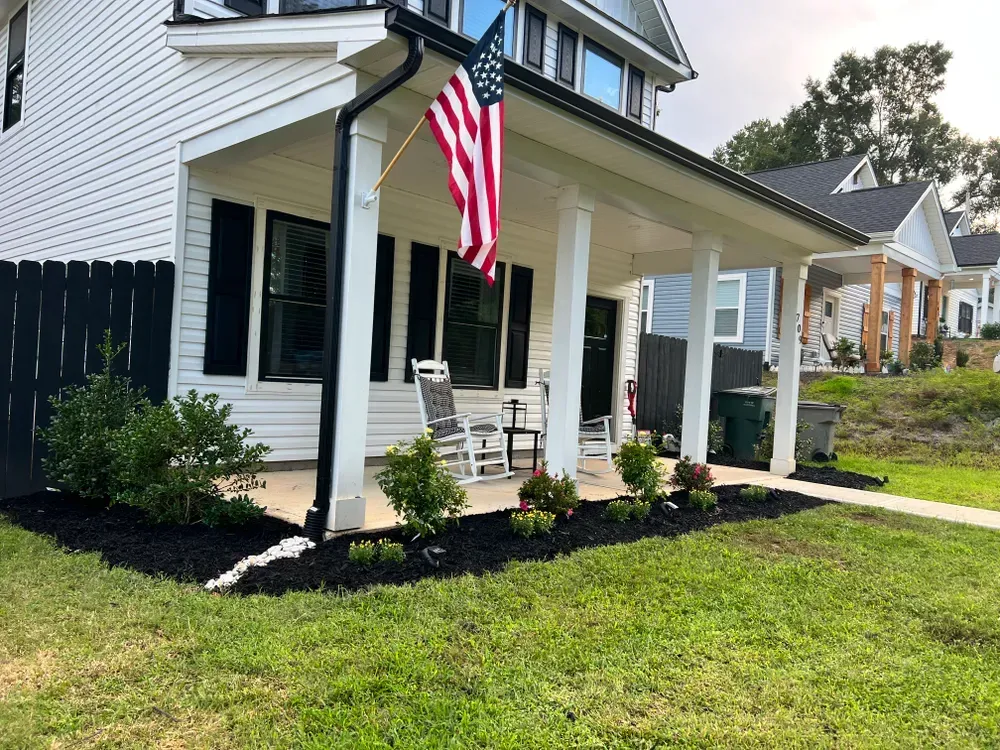 A white house with a covered porch, American flag, rocking chairs, and landscaped flower beds with black mulch.
