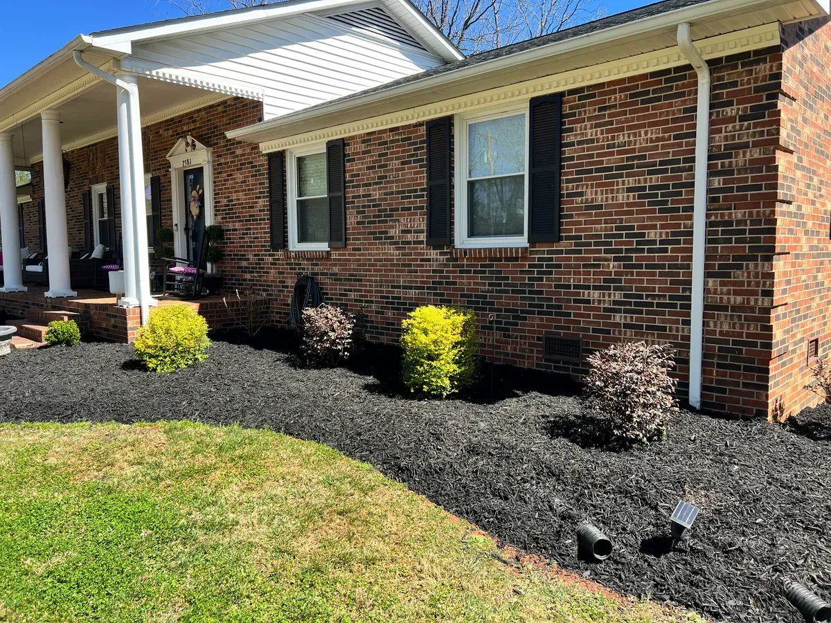 A brick ranch-style house with black shutters, white columns, and a landscaped front garden with dark mulch and shrubs.