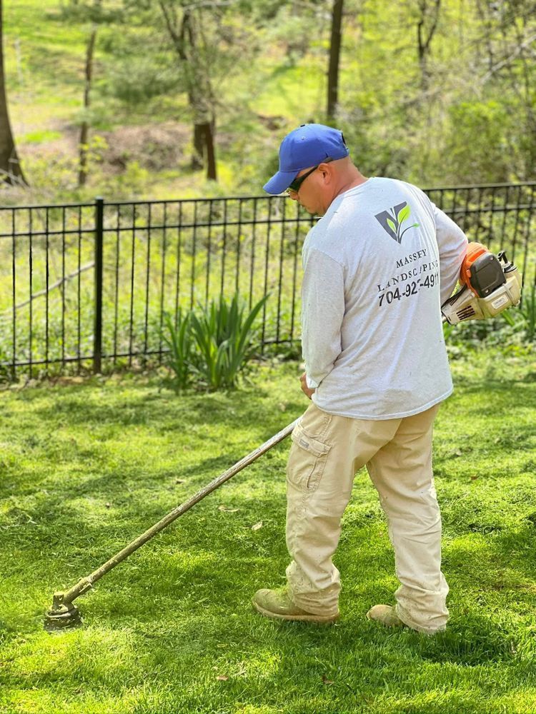 A worker in a blue cap and light-colored uniform uses a string trimmer to cut grass near a black metal fence.