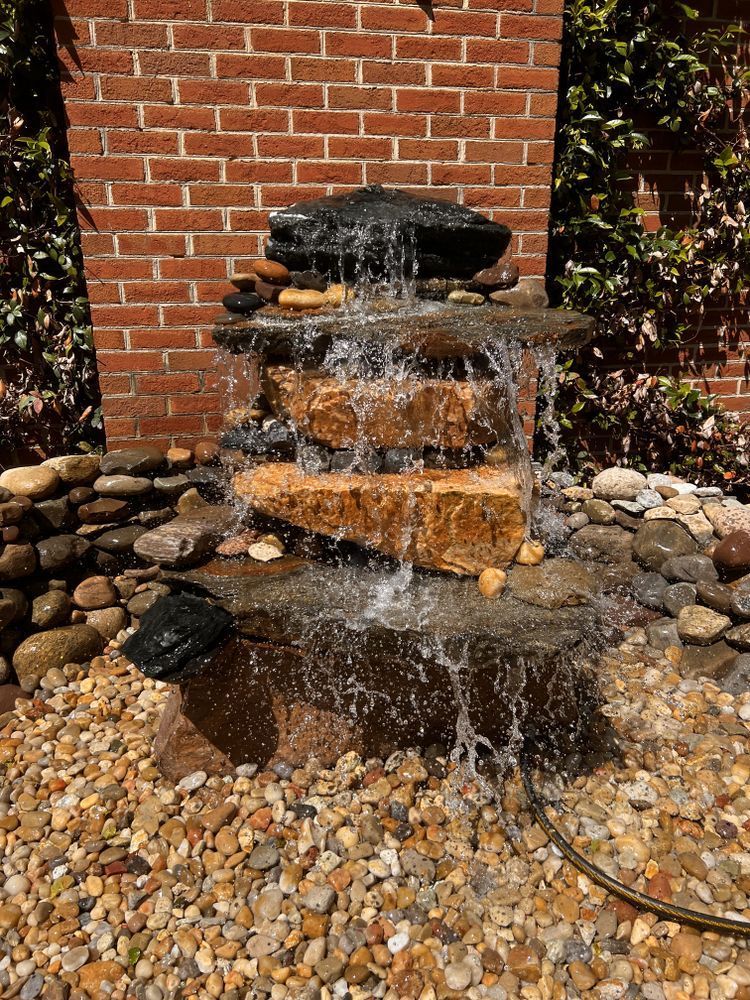 A tiered stone water fountain in front of a brick wall, with water cascading down layers into a bed of pebbles.