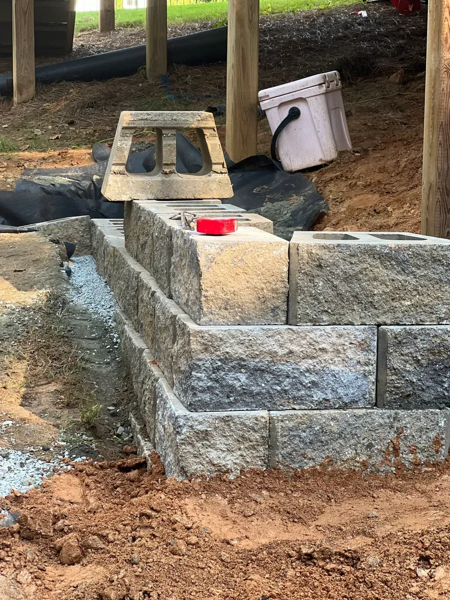 A partially built gray stone retaining wall under a wooden deck, with a single capstone and a red tape measure on top.