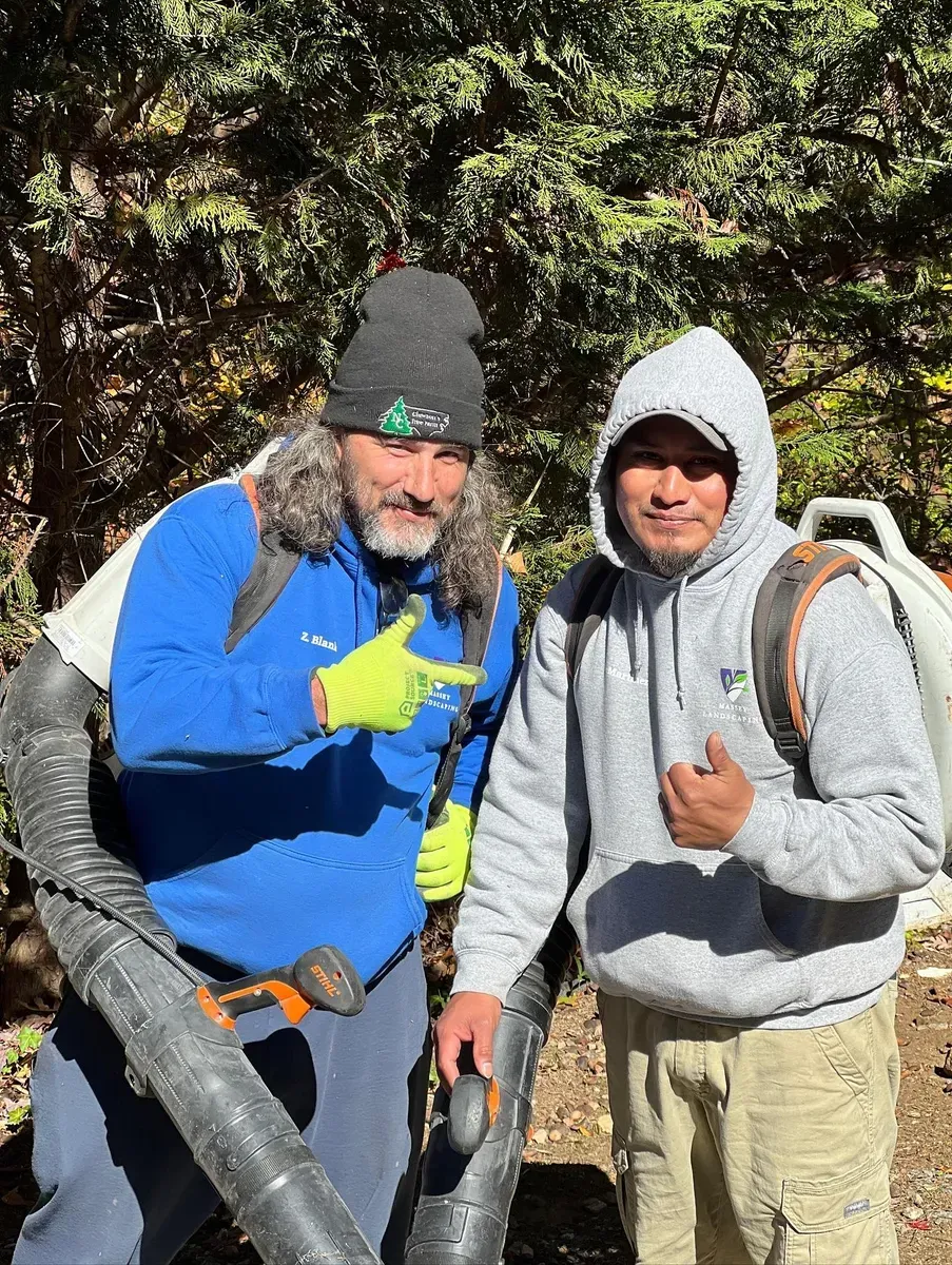 Two people stand in a wooded area wearing backpacks and holding leaf blower equipment while making hand gestures.