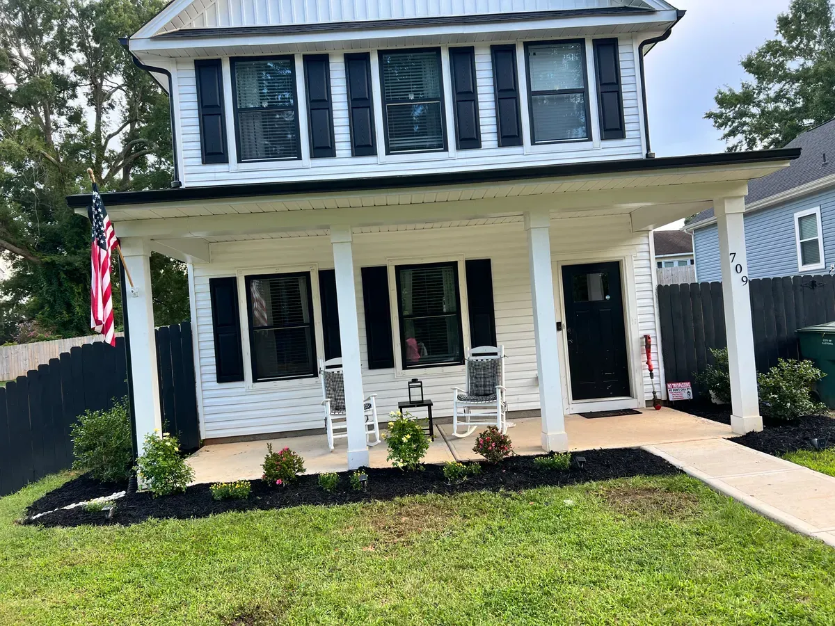 A two-story white house with black shutters, a front porch with two rocking chairs, and an American flag on the side.