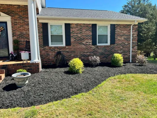A brick home exterior with black shutters, a white porch column, and a fresh black mulch bed featuring small green shrubs.