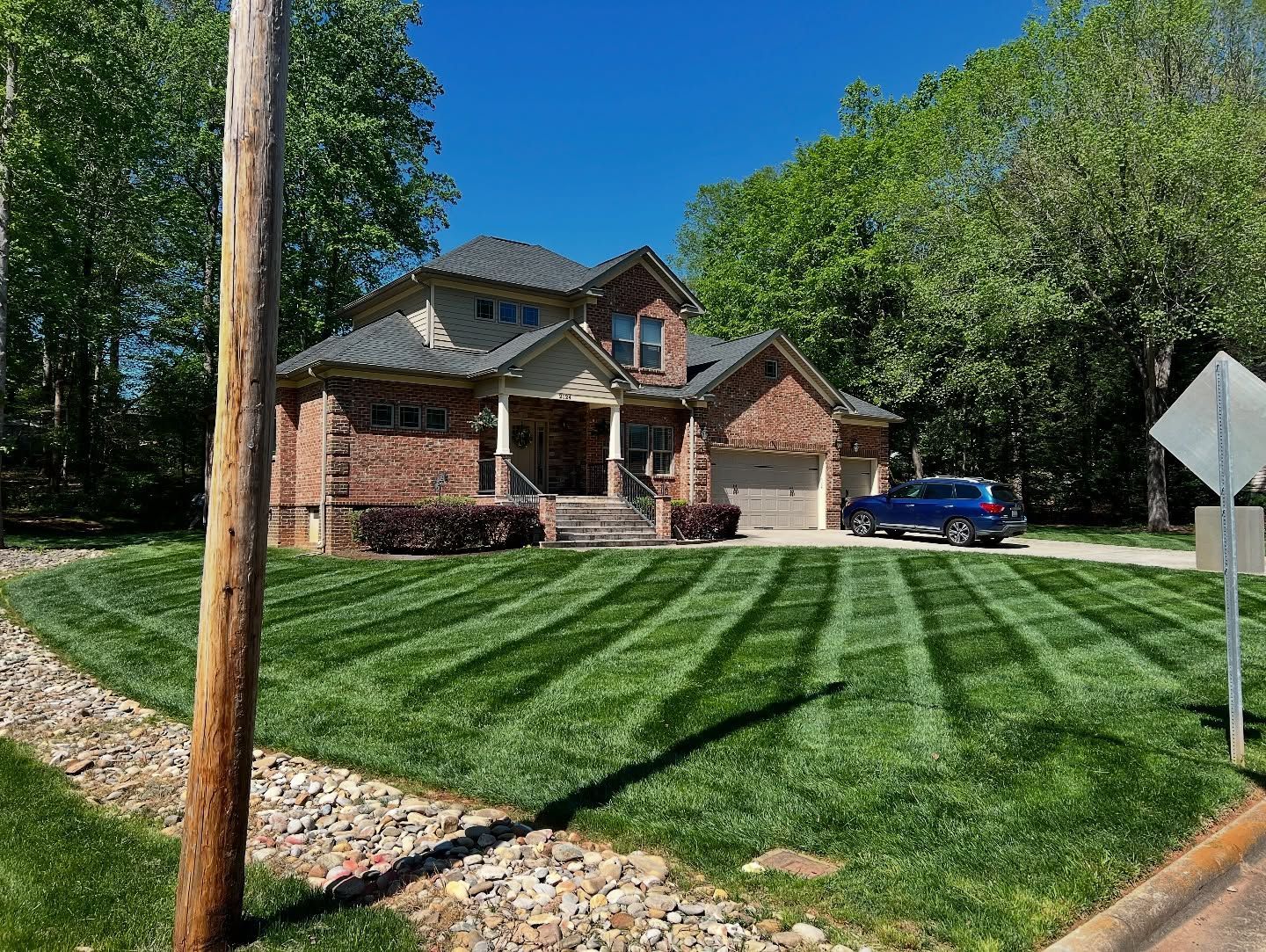 A two-story brick house with a manicured, striped green lawn and a blue car parked in the driveway on a sunny day.