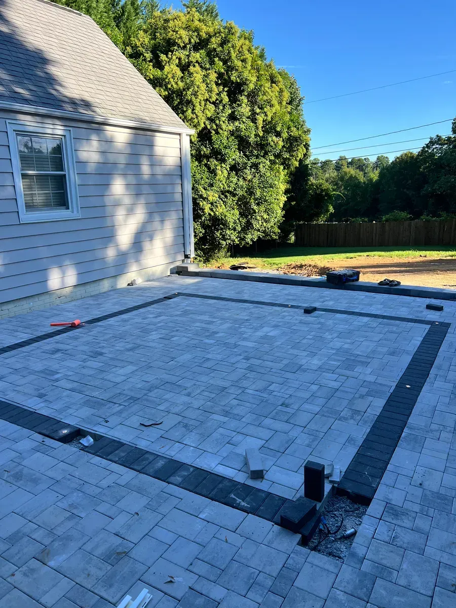 A newly installed gray paver patio with a dark border, attached to the side of a light-colored house near a grassy yard.